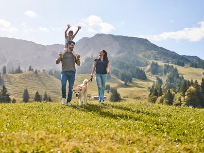 Alpin Chalets Oberjoch | Erlebnisse zwischen Action & Erholung Familie mit Hund wandert durch eine alpine Wiese vor Bergpanorama