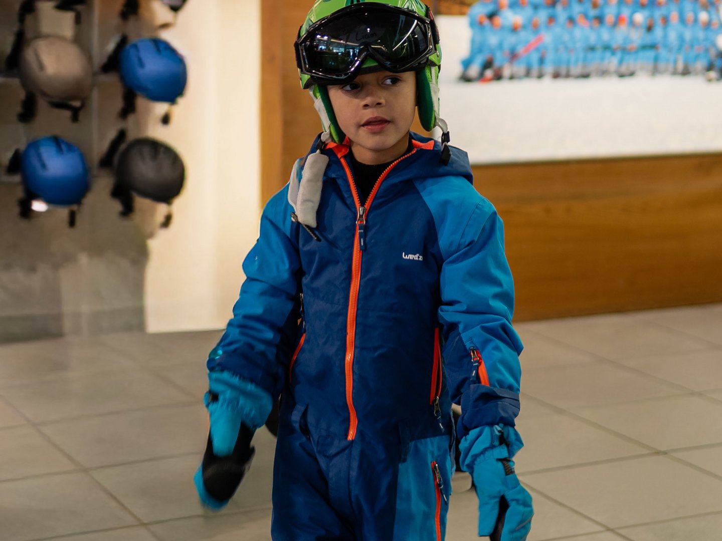 Skifahren lernen im Herzen des Allgäus Kind in blauem Skianzug, Helm und Skibrille neben einer Wand mit ausgestellten Helmen